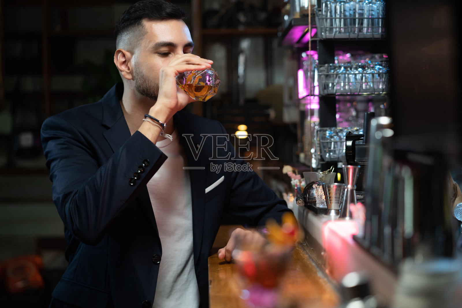 肖像英俊的白人男子坐在吧台拿着威士忌杯与冰，享受从酒吧的酒保喝美味的酒精饮料。夜生活派对庆祝和假日概念照片摄影图片