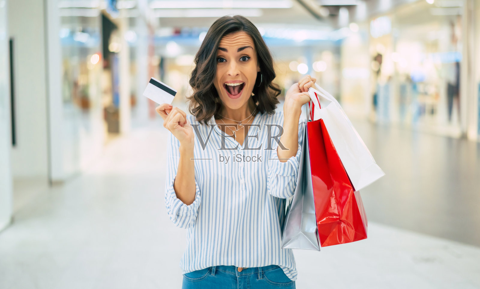 兴奋的美丽微笑的年轻女子是摆姿势与信用卡和购物袋在商场照片摄影图片