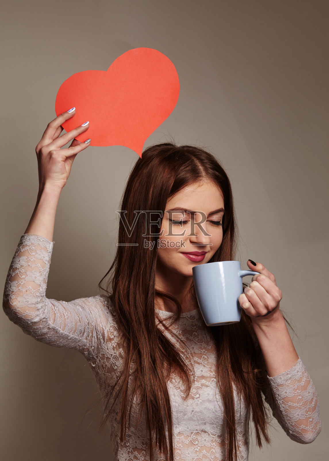 美丽的年轻女子在早晨爱上咖啡对着镜头微笑，在灰色的背景上，她手里拿着一个小心脏照片摄影图片