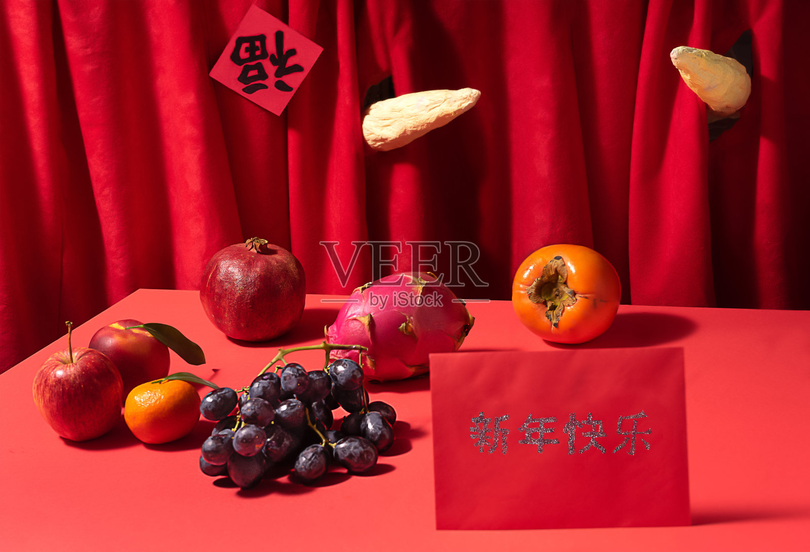两个牛角穿过深红色的背景。桌上摆着各种新鲜的水果。幕布上用粤语写着“好运”。红色的桌子上放着红色的幸运信封，上面写着“新年快乐”。照片摄影图片