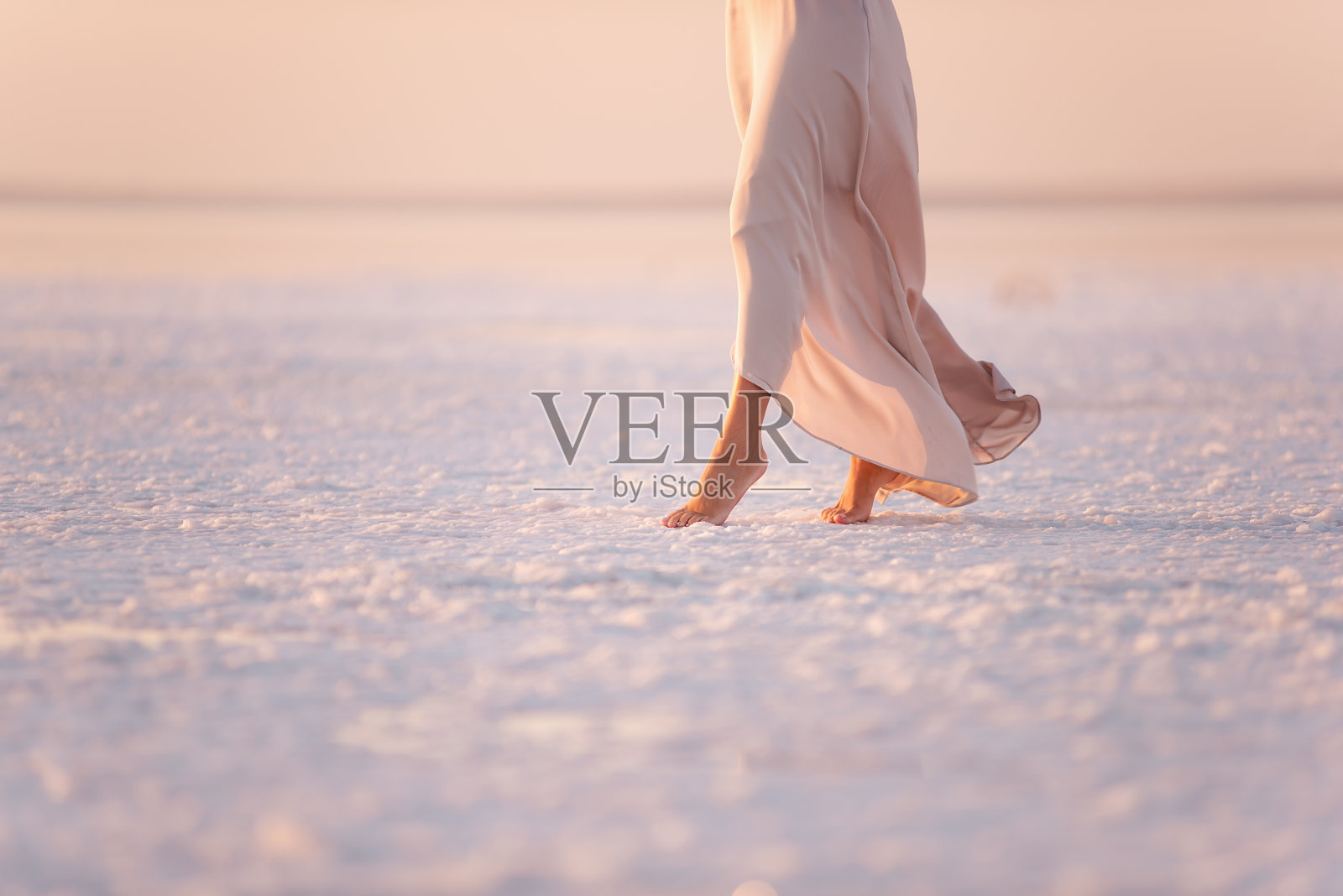 漂亮的整洁的腿年轻女子在傍晚通风柔和的粉红色连衣裙是走在水晶化的白色盐对日落的背景。矿物结构在河口提取有机盐照片摄影图片
