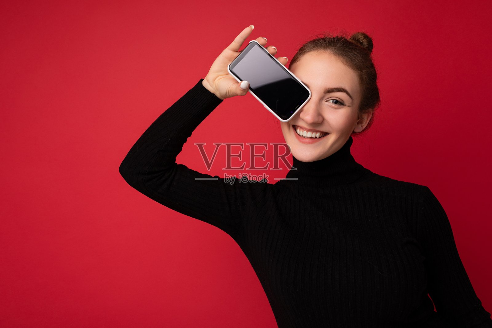 美丽的照片快乐微笑年轻的黑发女人穿着黑色毛衣站在孤立的红色背景显示手机空屏幕看模型的相机照片摄影图片