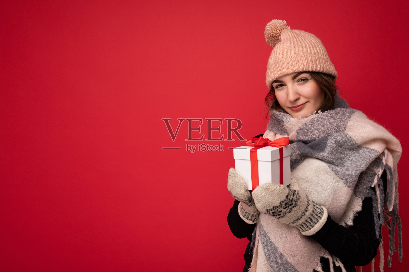 迷人的幸福可爱的年轻褐发女人孤立在红色背景墙戴着冬季围巾帽子和手套拿着白色礼盒与红色丝带和看着相机的舒适的镜头照片摄影图片