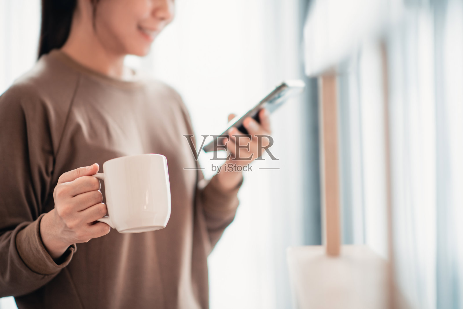 女人在家里用手机手捧一杯热茶或咖啡。照片摄影图片