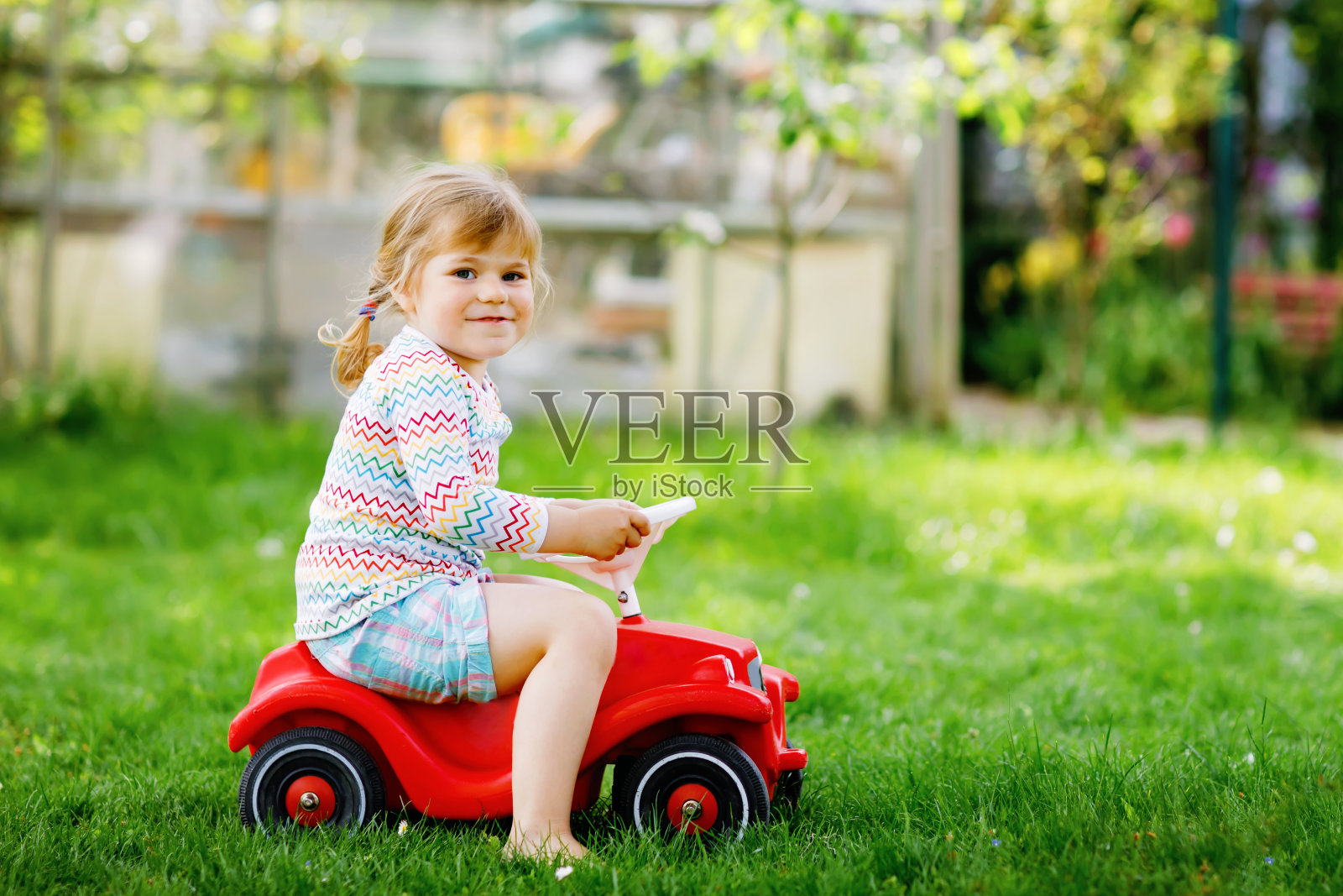 活泼可爱的小女孩开着玩具车，在户外玩得很开心。美丽快乐健康的孩子享受着温暖的夏日。在加登的迷人微笑的孩子照片摄影图片
