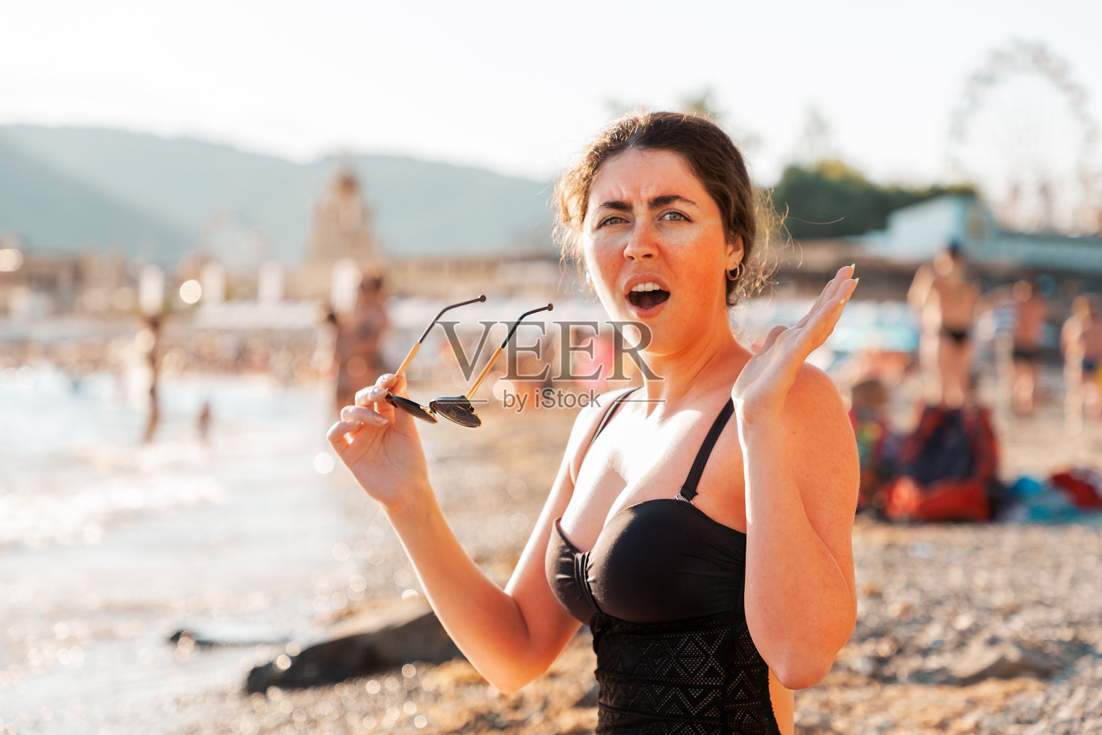 一个女人拿着太阳镜，因为晒伤而尖叫。面部和身体皮肤发红。背景是海滩和大海。保护和皮肤护理的概念照片摄影图片