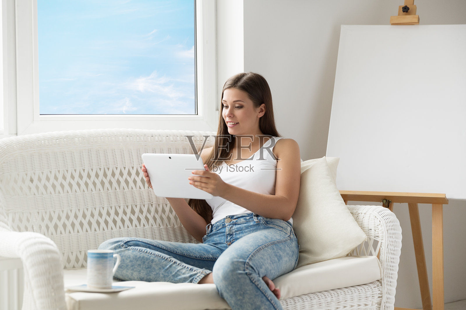 一个女人坐在沙发上看平板电脑，面前是空白的画架画布。小女孩用触控板屏幕休息在窗口旁边。在家用平板电脑的女人照片摄影图片