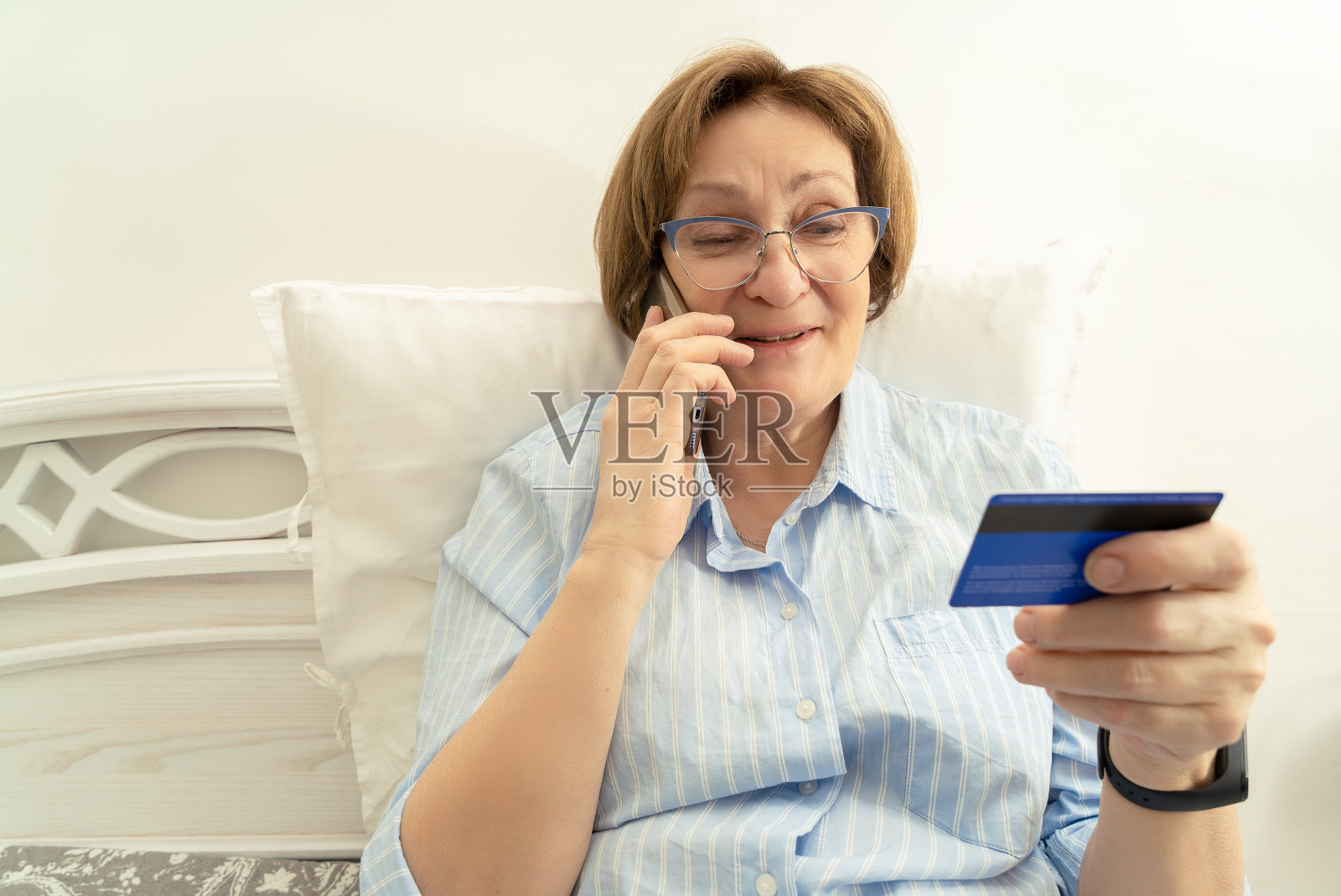 一位微笑的老妇人在室内用手机说话，手里拿着一张银行卡照片摄影图片