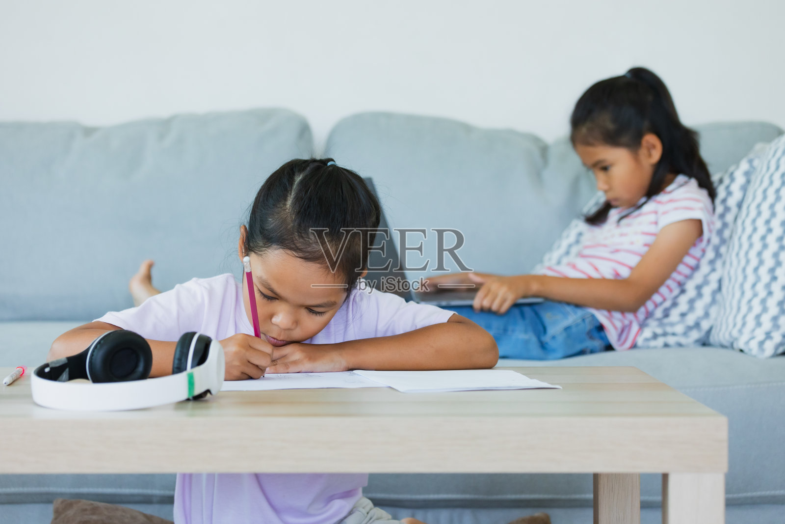 亚洲女孩做作业，她的妹妹坐在沙发上，并与老师和班级进行视频会议聊天。由于COVID-19疫情，儿童在隔离期间在家上学。照片摄影图片