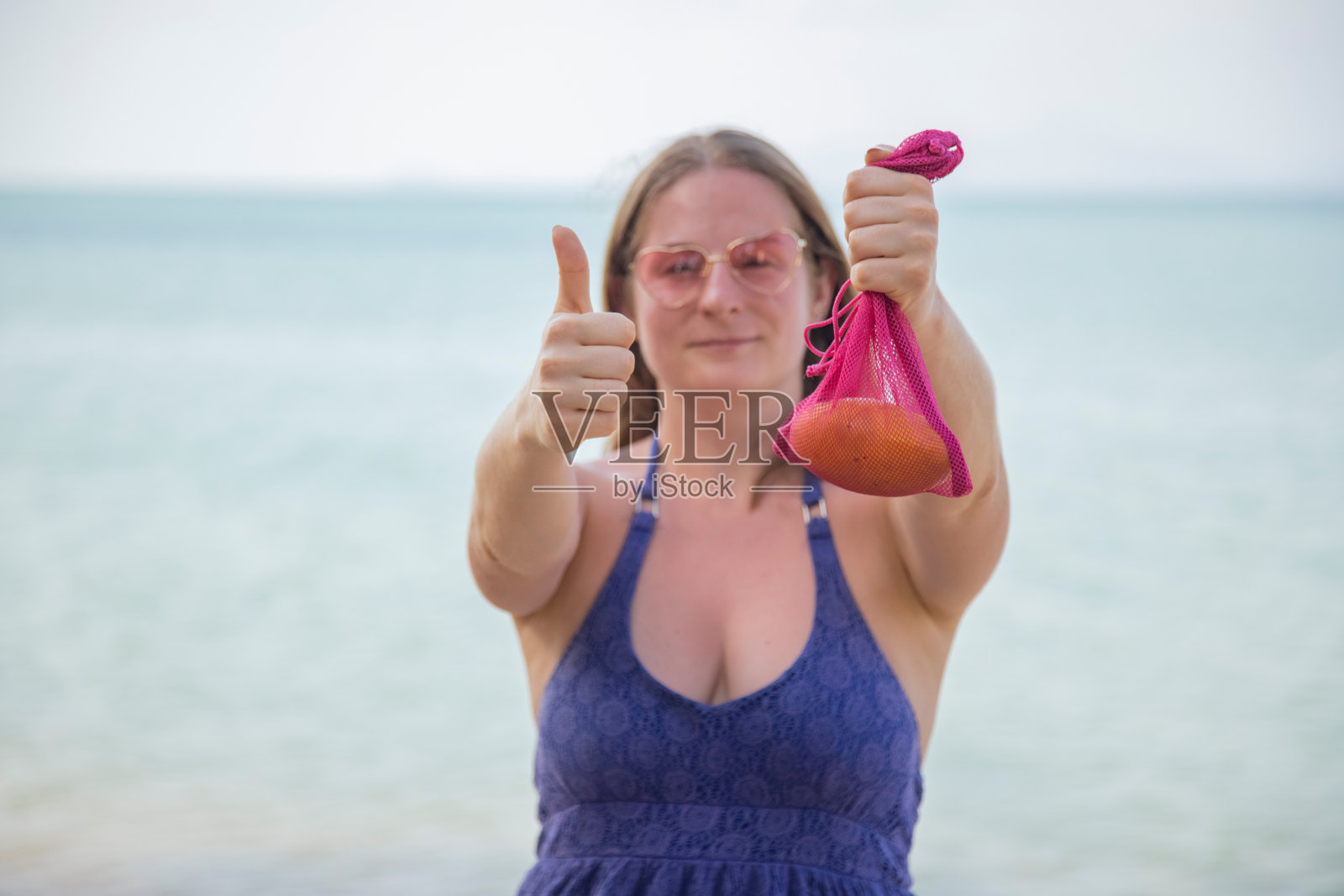 微笑的白人妇女拿着网袋水果和显示拇指向上。背景是大海或海洋。照片摄影图片