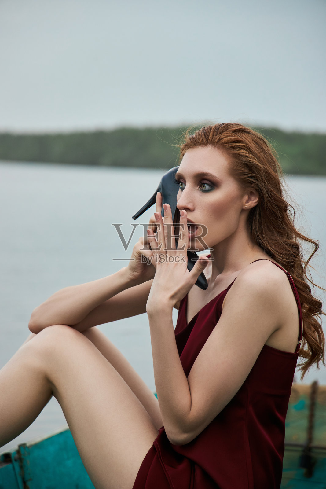 漂亮时尚的红发女人坐在船上。美丽浪漫的肖像女孩在湖上的木船上穿着红色葡萄酒的裙子照片摄影图片