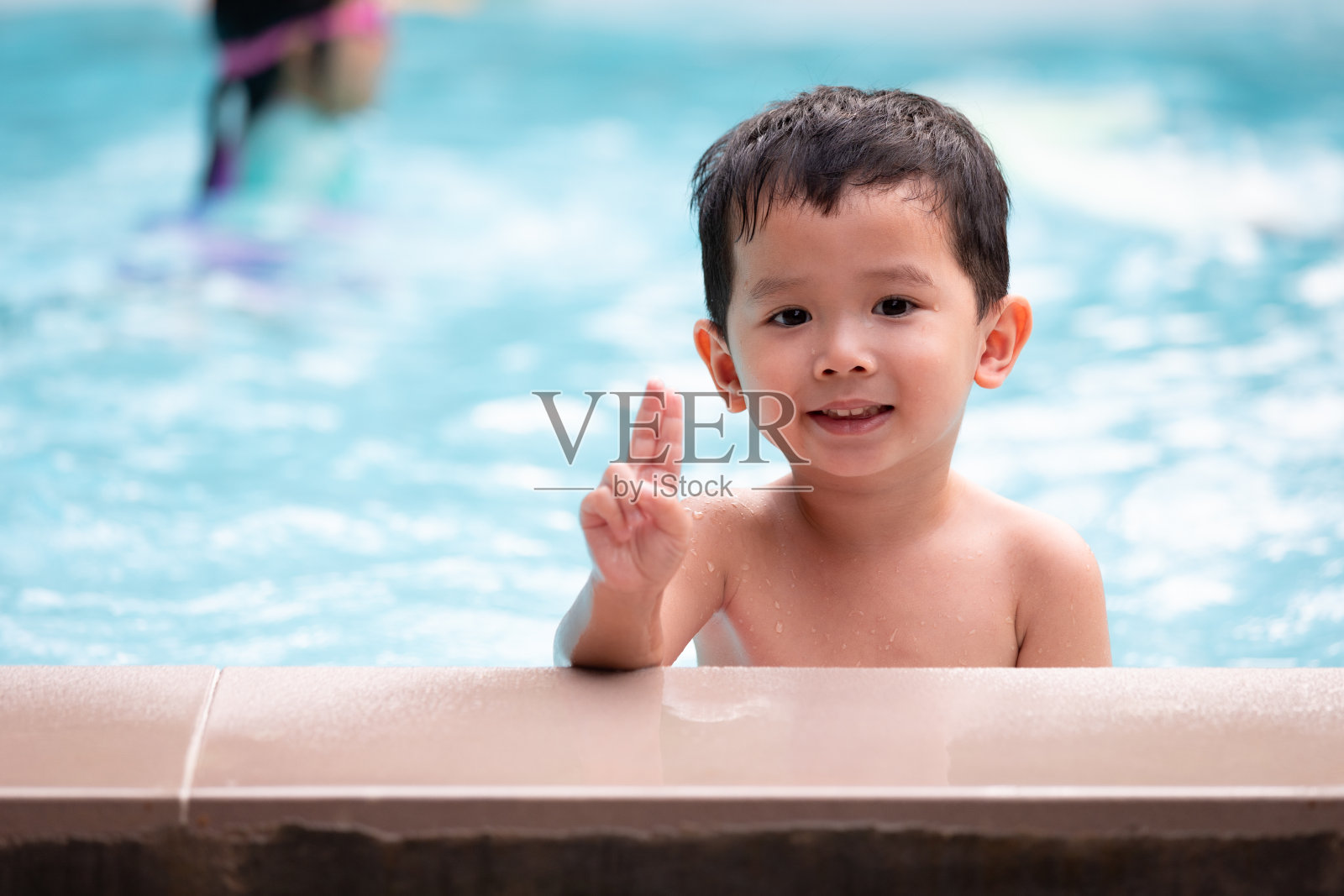 亚洲小男孩的肖像看着相机和微笑，而玩水在游泳池。照片摄影图片