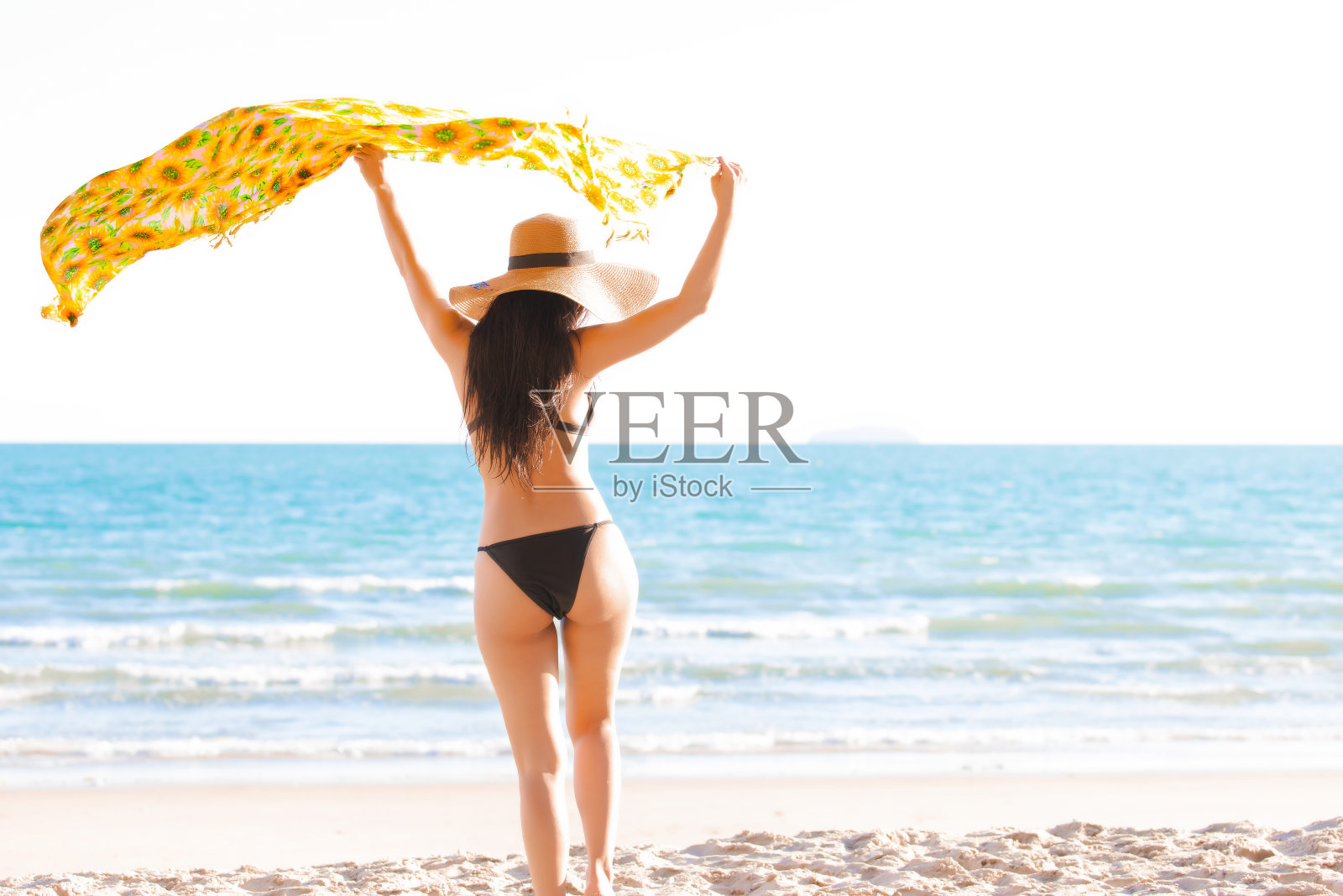 夏日里，热带岛屿的海滩上，穿着黑色比基尼的美女站在热带海滩上，手拿黄色围巾，戴着太阳帽。照片摄影图片