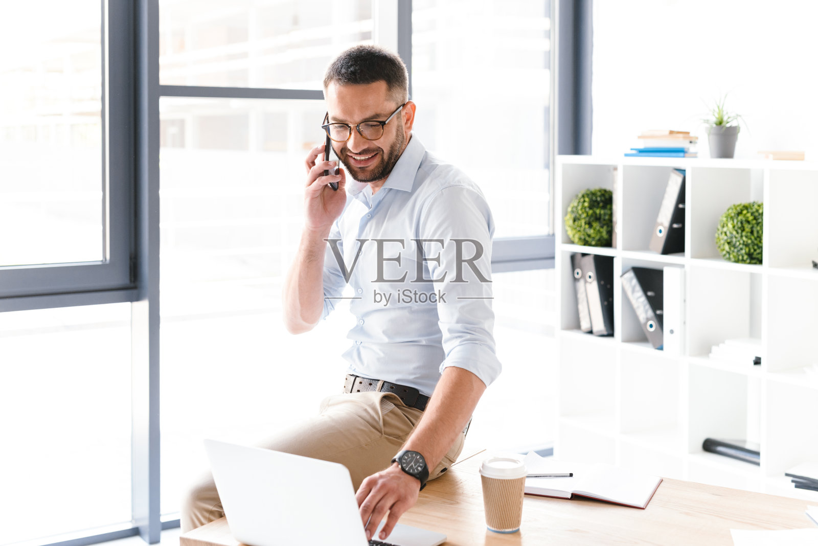 一个穿着正装的现代人坐在办公室的桌子上，用黑色智能手机谈生意照片摄影图片