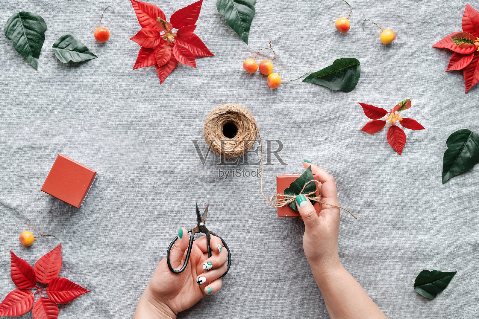 圣诞平铺在天然米色亚麻布上。手装饰礼品盒。红一品红，橘野苹果装饰。麻绳，剪刀，红色和绿色的一品红叶子照片摄影图片
