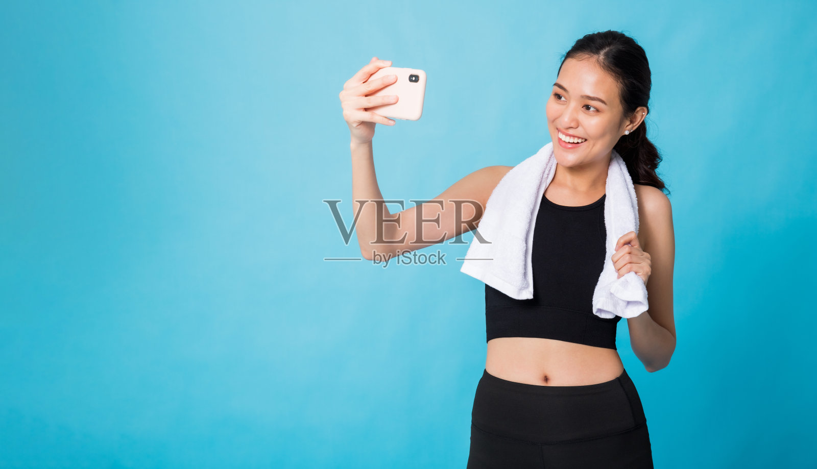 亚洲美丽快乐女子运动后手持智能手机自拍，隔离在蓝色背景下与摄影棚拍摄。概念苗条和健康的女孩锻炼。照片摄影图片