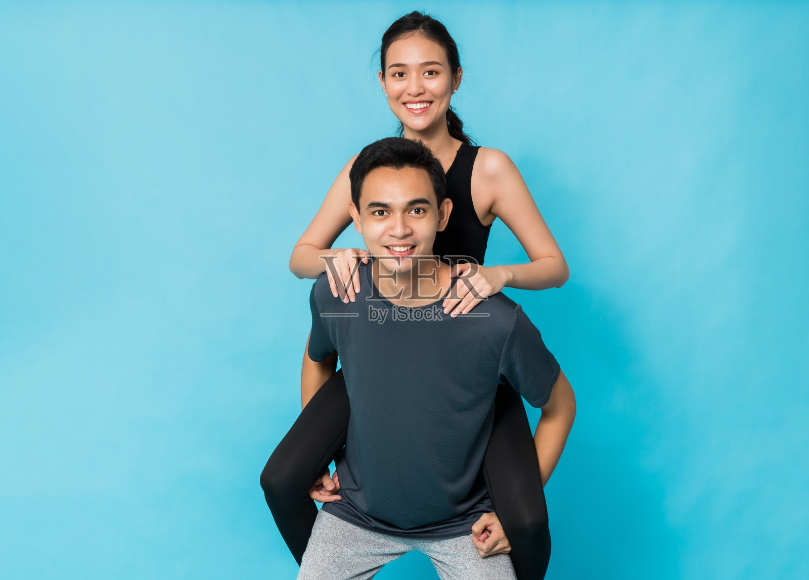 亚洲美丽快乐的女人骑在男朋友的背上运动后孤立在蓝色背景。概念苗条和健康的女孩锻炼。照片摄影图片