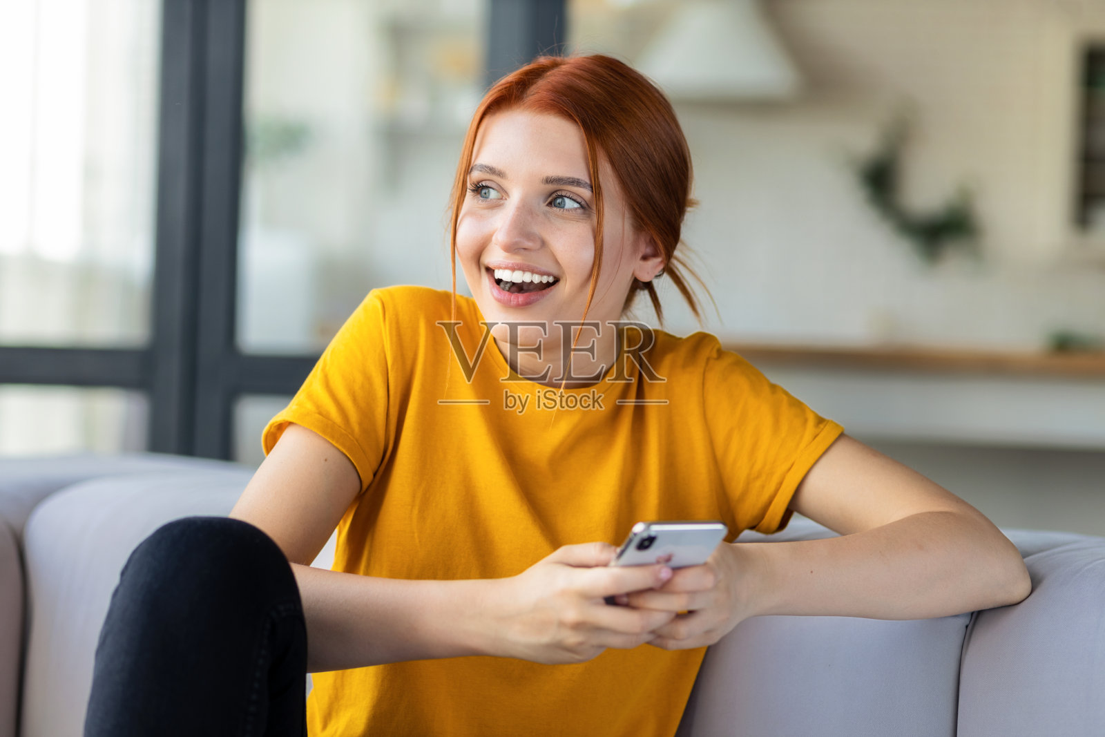 快乐的红发女孩在社交网络上发短信，坐在家里的沙发上用手机发短信。在线沟通，科技理念照片摄影图片