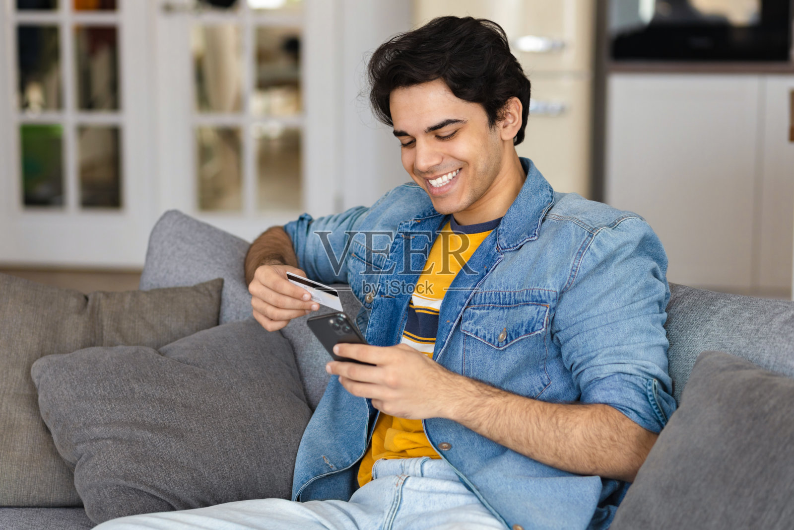 快乐微笑的年轻男或学生用信用卡使用手机在互联网上赚钱的购买坐在家里的沙发上照片摄影图片