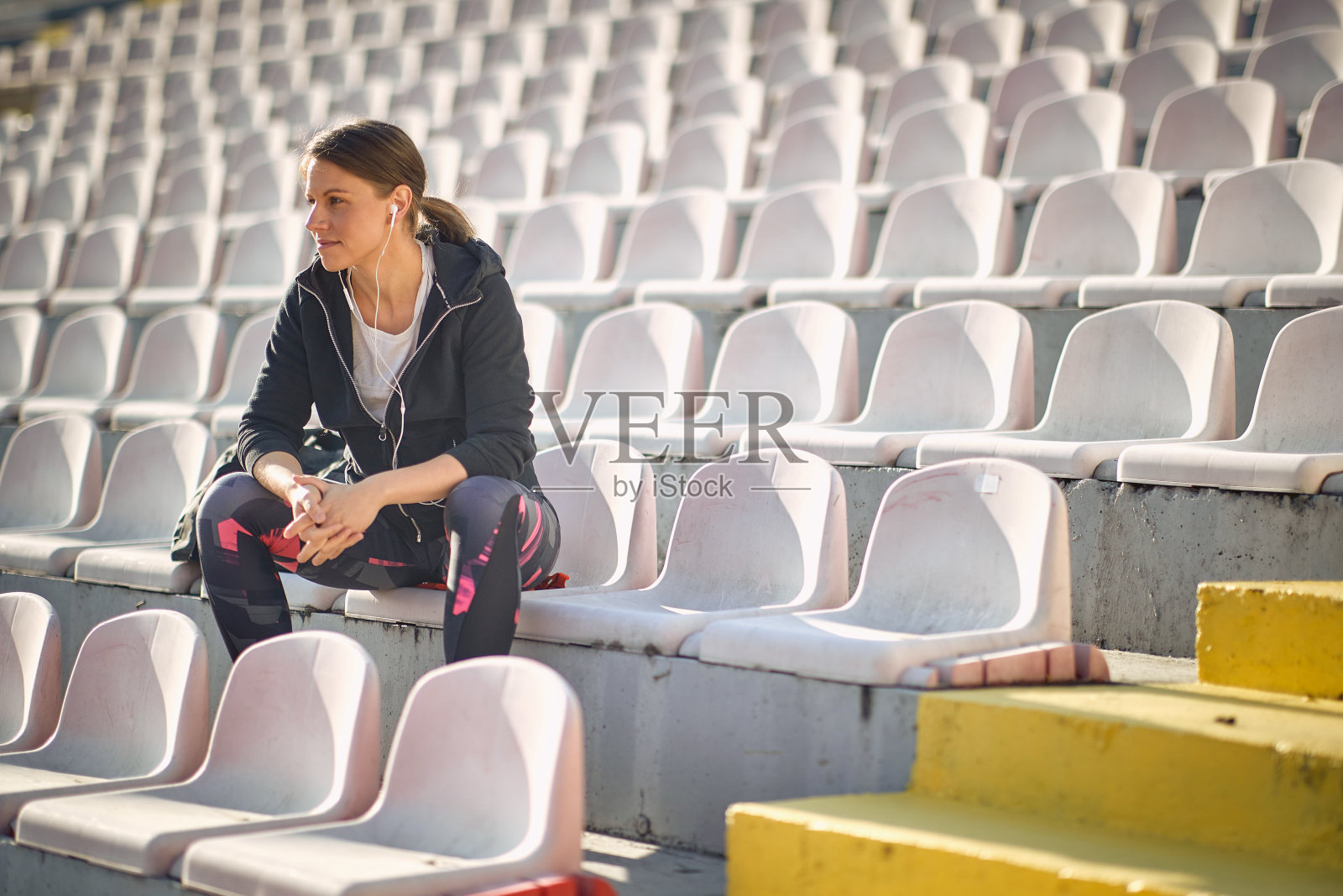 漂亮的年轻白人女性坐在体育场里。戴着耳机看着远方照片摄影图片