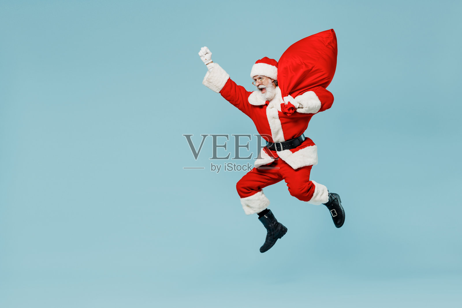 全身上下50多岁的圣诞老人戴着圣诞帽，穿着红色西装，像超级英雄一样跳着，带着礼物袋，孤立在素色背景的工作室里。新年快乐，2022年圣诞快乐照片摄影图片