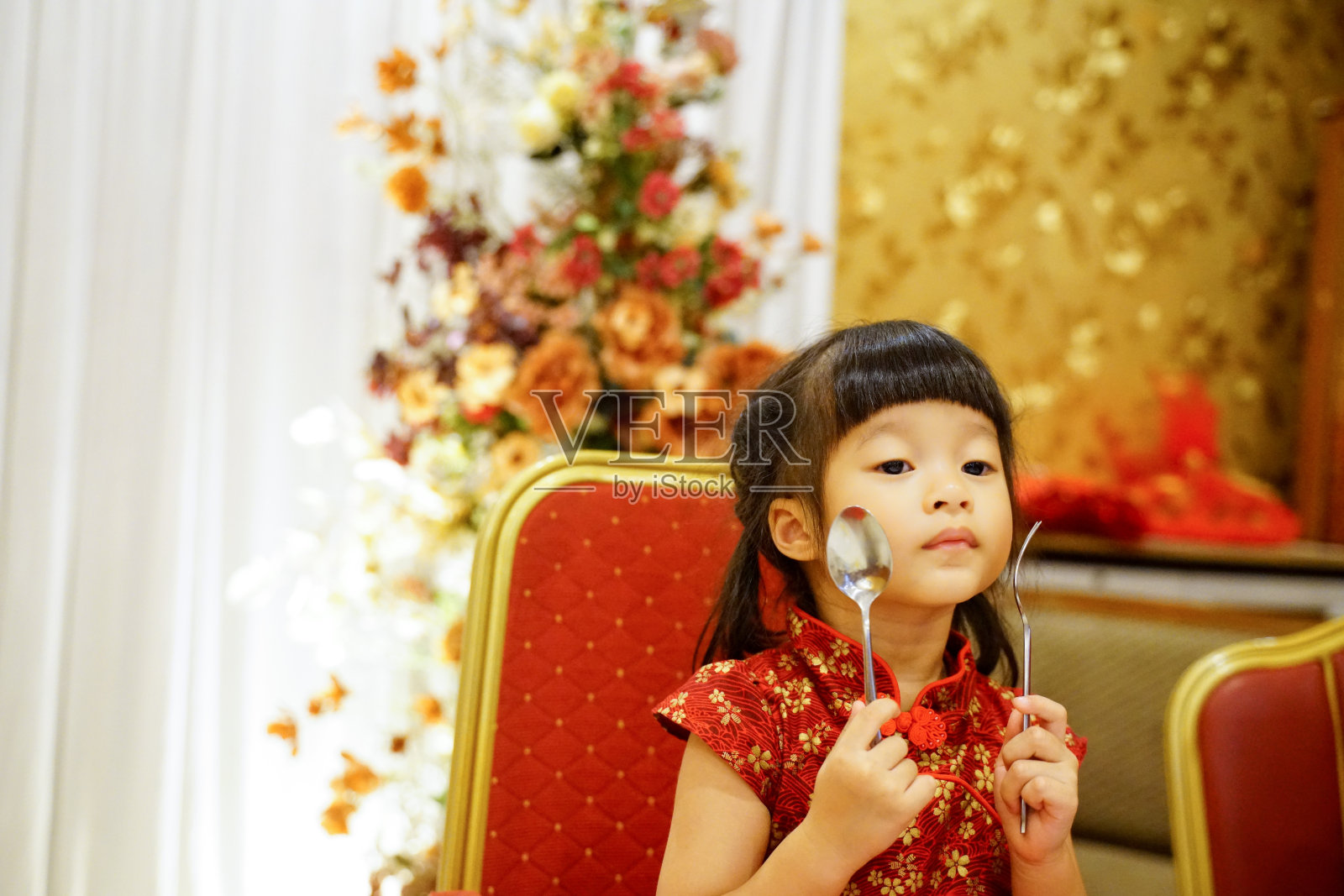 女婴拿着叉子和勺子秀在脸上感到高兴的春节晚会期间，孩子在餐厅里穿着红色的服装坐在椅子上。照片摄影图片